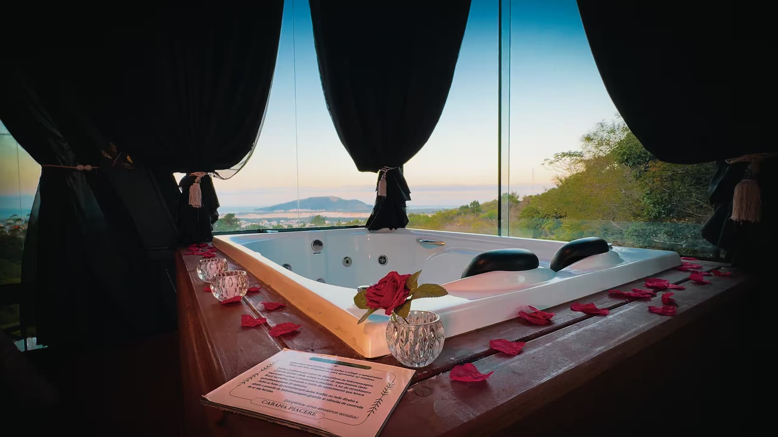 Banheira de hidromassagem decorada com pétalas de rosas, diante da janela com vista para o mar ao entardecer